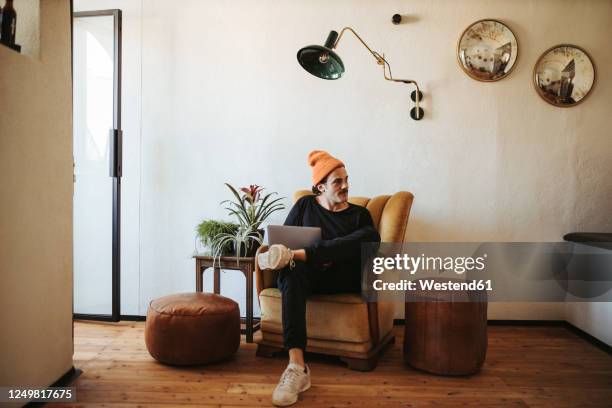 man with laptop sitting on armchair at home - homme hipster stock-fotos und bilder