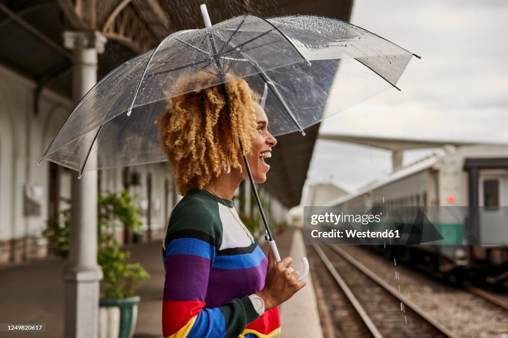 Side view of cheerful young woman enjoying while standing with umbrella at railroad station during monsoon