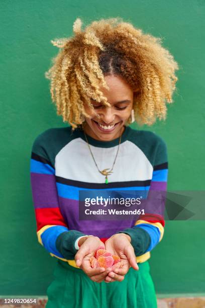happy young woman with afro hairstyle holding candy hearts against green wall - holle handen stockfoto's en -beelden