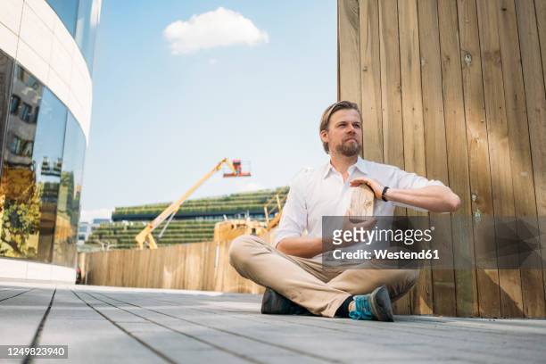 businessman sitting on the ground at construction site holding wooden house model - architekturmodell stock-fotos und bilder