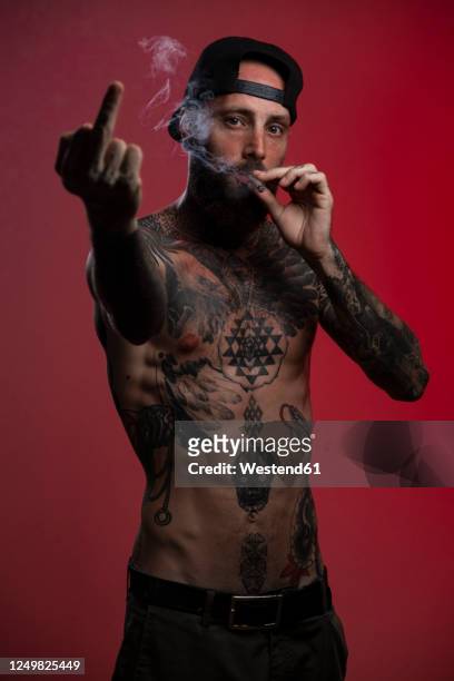 portrait of tattooed man against red background giving the finger while smoking a joint - marijuana tattoo stock pictures, royalty-free photos & images