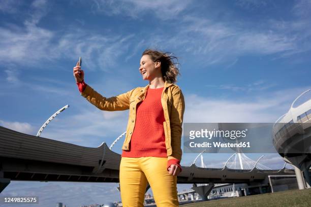 happy woman with windswept hair taking a selfie at a road bridge - yellow coat stock-fotos und bilder