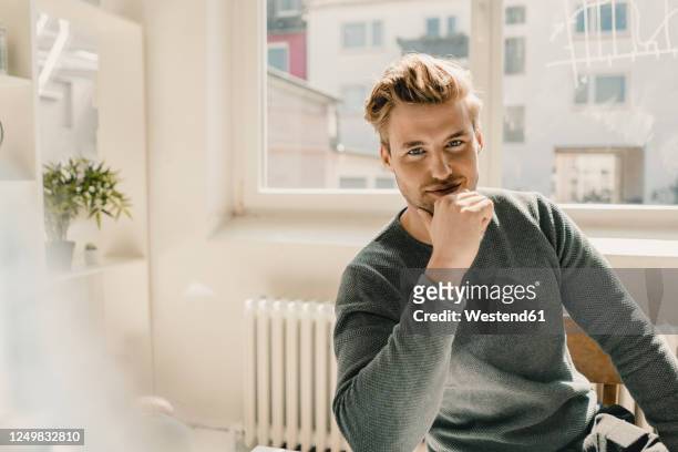 smiling young man sitting in office, hand on chin - lässige kleidung stock-fotos und bilder