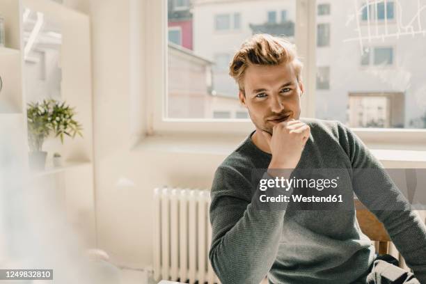 smiling young man sitting in office, hand on chin - man hand on chin stock pictures, royalty-free photos & images