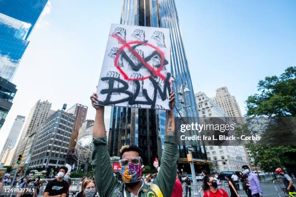 Protester wearing a mask holds a homemade sign above their head that says, "BLM" with a swastika in a circle with a line through it and a pattern of...