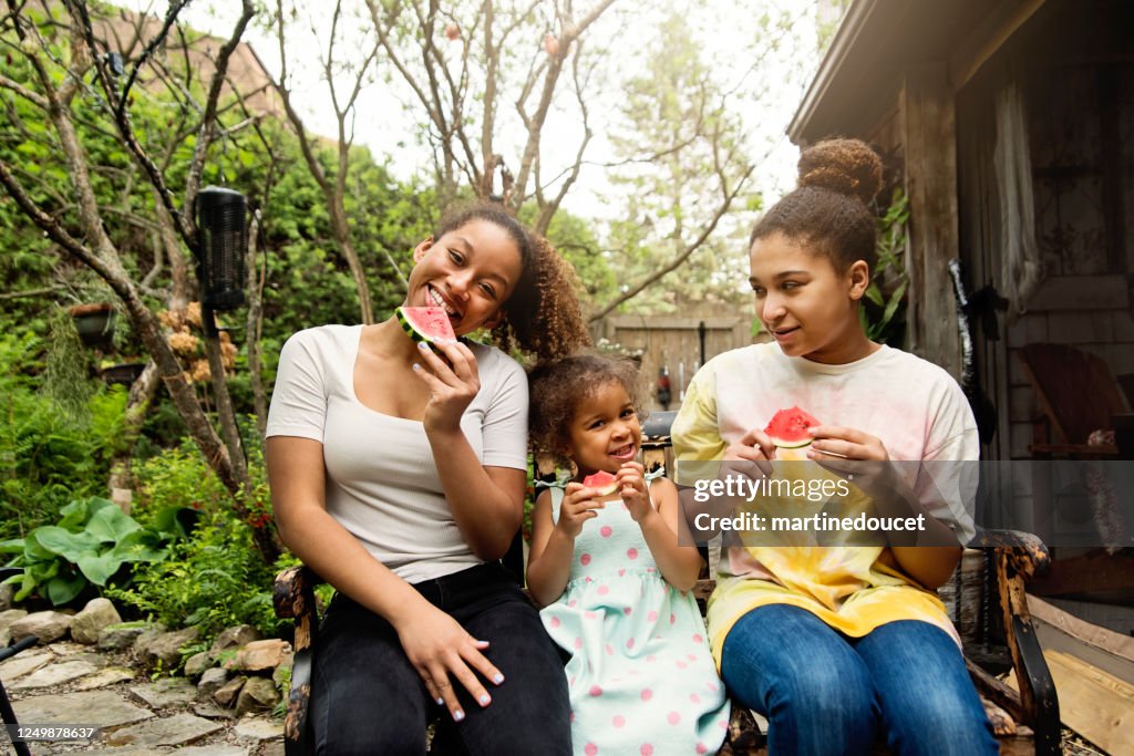 Mixed-race sisters eating watermelon in backyard.