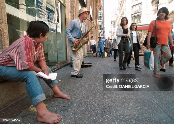 Ciudadanos argentinos pasan frente a un hombre que mendiga tocando el saxo en la peatonal Florida el 05 de Diciembre mientras un nino descalzo...