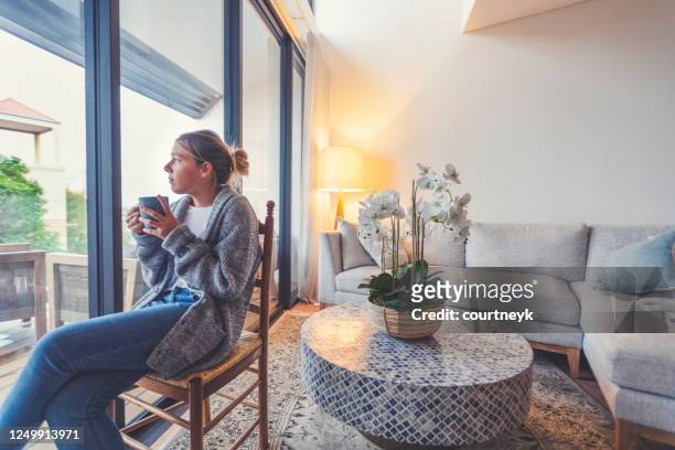 woman looking through the window drinking tea or coffee. - lockdown loneliness stock pictures, royalty-free photos & images