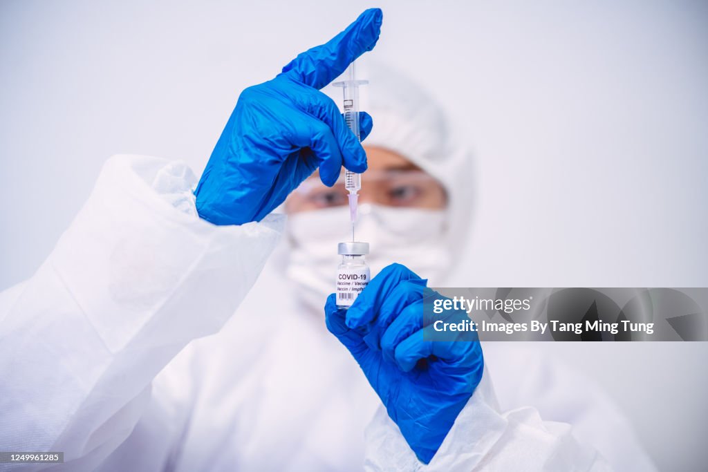 Doctor in protective gloves & workwear filling injection syringe with COVID-19 vaccine