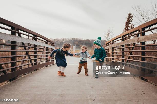 three siblings laughing holding hands running on bridge - family sports centre laughing stock-fotos und bilder