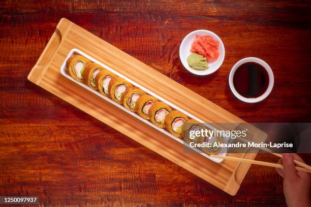 woman's hand holding sticks to eat an asian meal consisting of sushi with crab and avocado stuffing, banana tempura around rice accompanied by soya sauce, pickled ginger and wasabi on a wooden table - sushi plate stock pictures, royalty-free photos & images