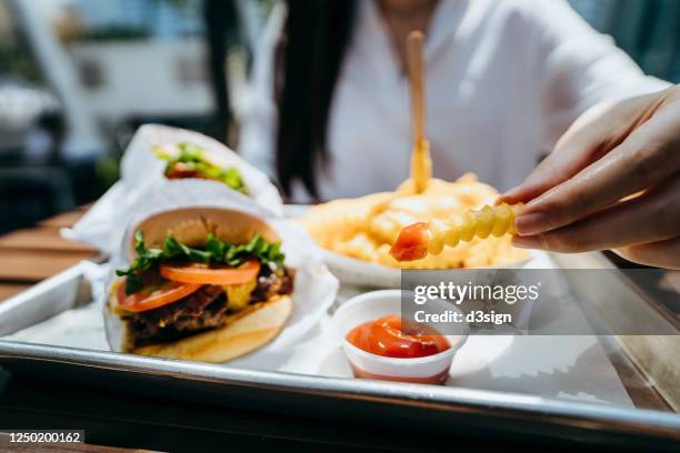 close up of young asian woman dipping fries with ketchup and eating beef burger in an outdoor fast food restaurant - snabbmatsrestaurang bildbanksfoton och bilder