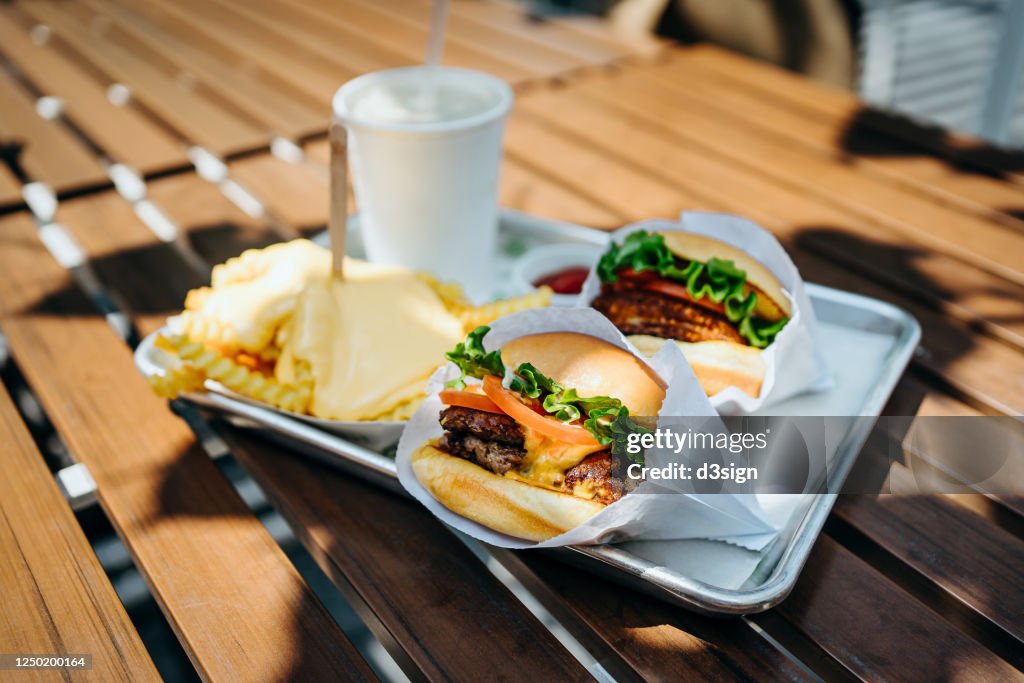 Freshly made beef burgers with cheese fries and soft drink on a tray served on a table with sunlight in an outdoor fast food restaurant