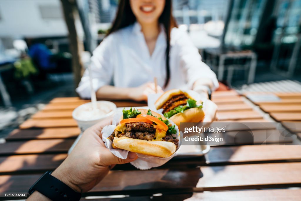 Happy young Asian couple eating beef burger with fries in an outdoor fast food restaurant