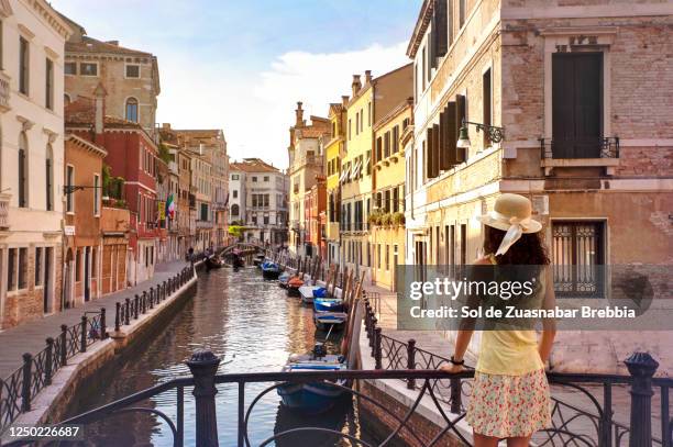 pretty woman wearing hat, yellow t-shirt and matching skirt standing on a small venice bridge looking down the canal of the venice lagoon - gondola traditional boat stockfoto's en -beelden