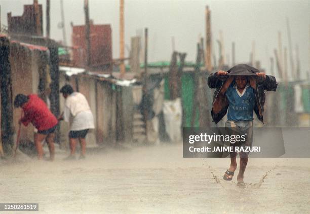 Child shields himself from torrential rains with his jacket as he runs through a flooded street on March 17, 1998 while local residents sweep mud and...