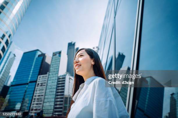 low angle view of confidence young asian businesswoman looking away while standing outside corporate building in urban downtown financial district. city scene in background with natural sunlight - low motivation stock pictures, royalty-free photos & images