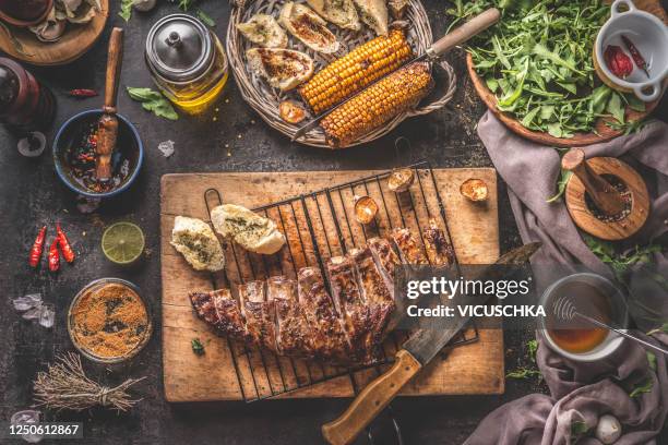 tasty grilled bbq meat ribs on wooden cutting board and grill grate with knife and fresh garlic herb butter bread served on rustic table with grilled corn cobs with bbq sauce , salad leaves and other tasty ingredients - costeleta comida imagens e fotografias de stock