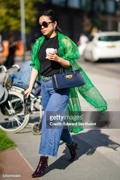 Guest wears sunglasses, a green mesh long vest, a Prada leather bag, a black t-shirt, a belt, blue flared ripped denim jeans, burgundy leather pointy...