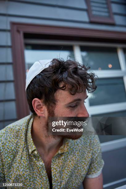 a young jewish man sitting at a table outside wearing a yamika, looking off camera. - jewish man fotografías e imágenes de stock