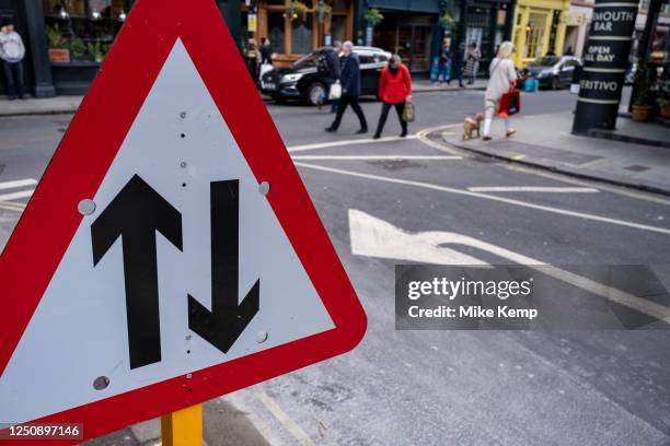 Two-way street traffic straight ahead sign interacts with passing people and an arrow painted on the road on 27th March 2023 in London, United...