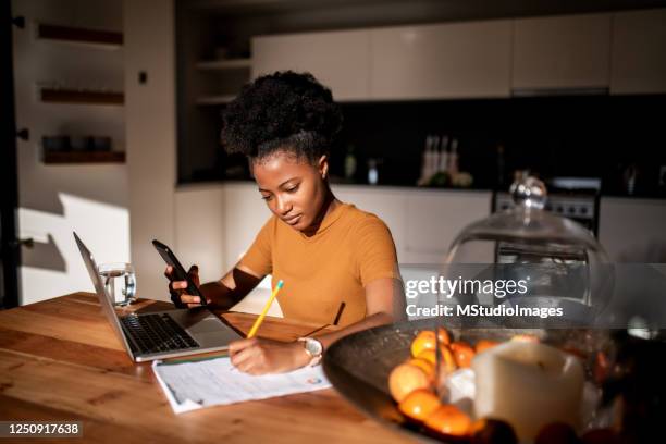 young woman learning online with laptop and phone, she is taking notes in a notebook - kitchen sunlight stock pictures, royalty-free photos & images