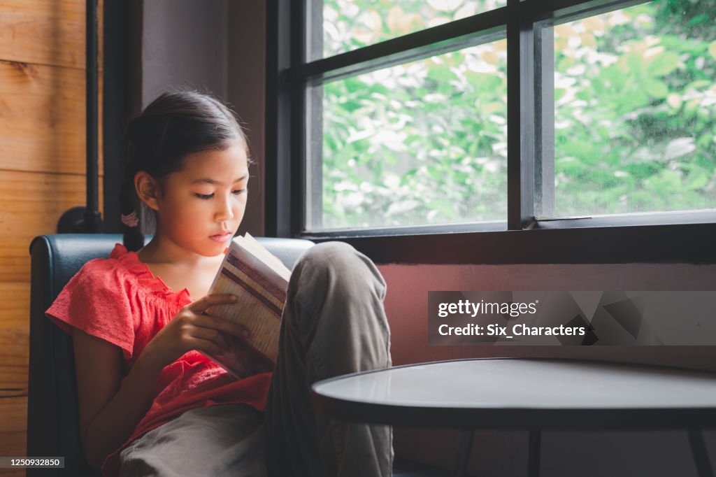 Cute little Asian girl reading a book in the living room at home, Education and knowledge concept