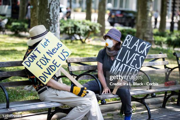 Two protesters wearing masks and sitting on a park bench observing social distancing hold homemade signs that say, "Don't Let Fox News Divide Us"...