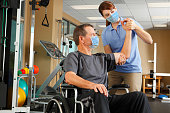 Physical Therapist And Patient In Wheelchair Wearing Protective Masks While Therapist Evaluates Range Of Motion