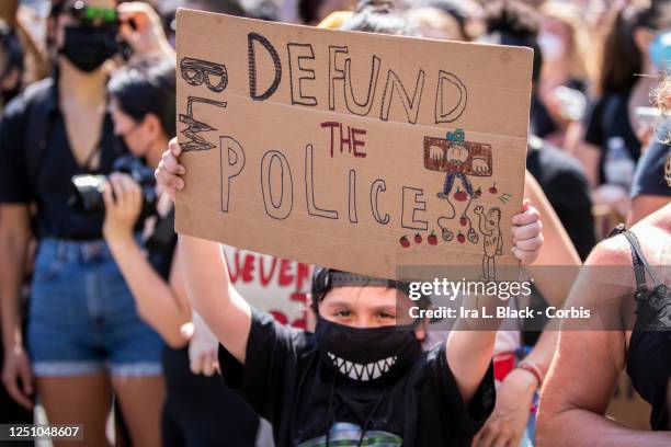 Young protester wearing a mask holds a homemade sign that says, "Defund The Police" with a hand drawn picture of a police officer held in stockades...