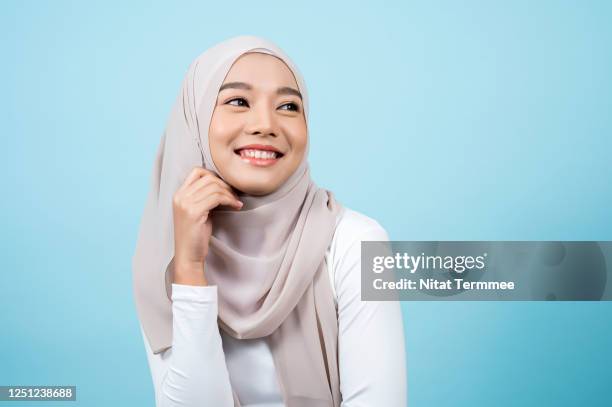 smiling asian muslim woman wearing headscarf while standing with a light blue background. muslim people concepts - asian model stock pictures, royalty-free photos & images