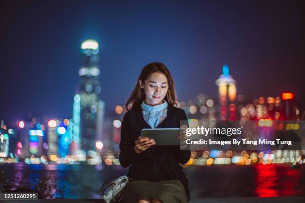 young cheerful asian woman using digital tablet at promenade against illuminated cityscape at night - asien e commerce stock-fotos und bilder