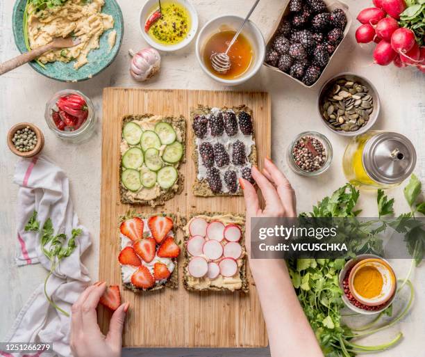 women hands making sandwiches with green healthy gluten free chips bread oder falafel bread with fresh herbs, chickpeas hummus , nuts and olive oil , berries and vegetables on white kitchen table with ingredients. - snacks stock-fotos und bilder