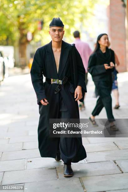 Guest wears a hat, a black long kimono outfit with flared pants, a belt, black leather shoes, during London Fashion Week September 2019 on September...