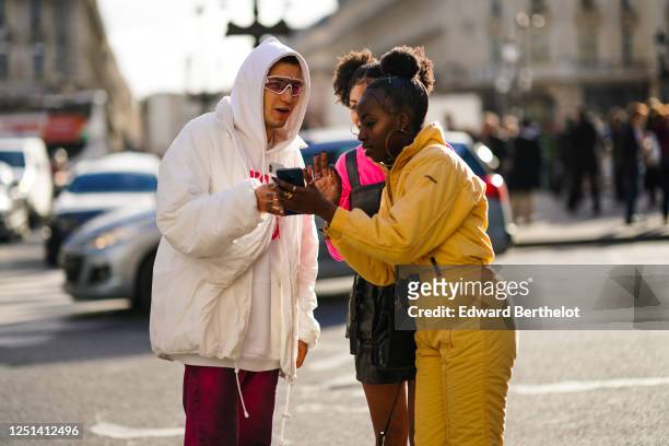 Guest wears pink ski goggles, a white hooded sweatshirt, a white padded jacket, burgundy pants ; A guest wears earrings, rings, a yellow ski suit,...