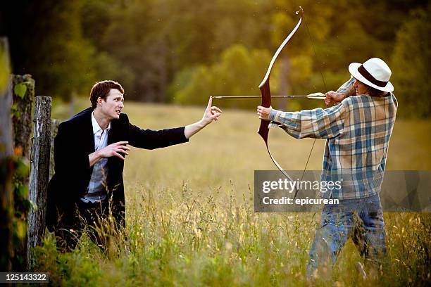 young man firing bow and arrow towards a businessman - bow arrow stock pictures, royalty-free photos & images