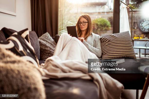 eine alleinstehende frau liest ein e-book auf ihrem digitalen tablet, während sie zu hause auf ihrem sofa liegt - e reader stock-fotos und bilder