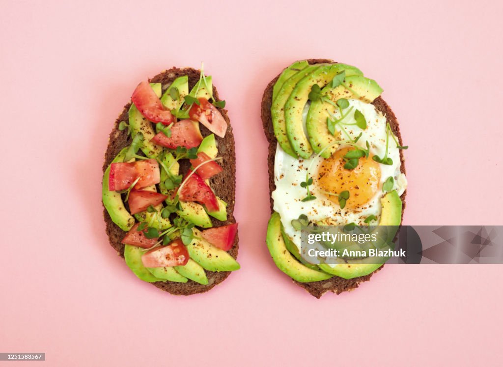 Toasts of dark bread with avocado slices, red tomatoes, fried egg and microgreen. Top view with pink background.