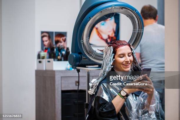 lächelnde junge frau mit handy während des wartens auf haarfärbemittel zu trocknen in friseursalon - at hairdresser stock-fotos und bilder