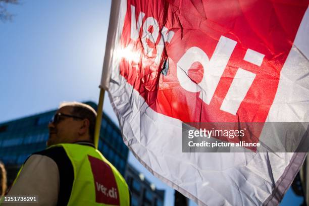 April 2023, Berlin: A participant of a rally on the occasion of the warning strike at Galeria, Ikea and Thalia holds a Verdi flag in front of the...