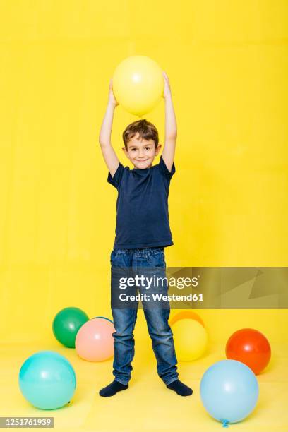 full length portrait of smiling boy holding balloon against yellow background - child balloon studio photos et images de collection