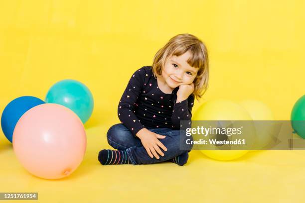 portrait of smiling cute girl sitting with colorful balloons against yellow background - child balloon studio photos et images de collection