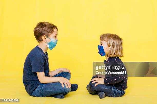 full length side view of siblings wearing face masks while sitting against yellow background - protective face mask side stock pictures, royalty-free photos & images