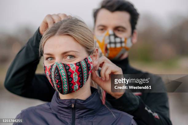 portrait of young woman while man adjusting face mask for her at park - epidemie stock-fotos und bilder
