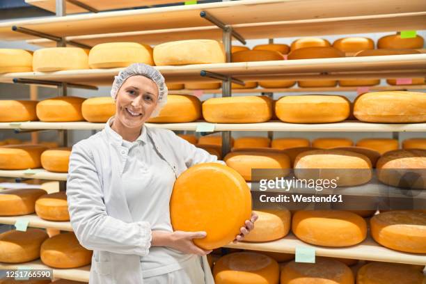 cheese factory, smiling female worker holding cheese wheel - käselaib stock-fotos und bilder