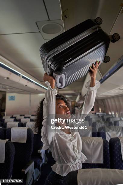 young woman positioning luggage inside storage compartment in airplane - positioned stock-fotos und bilder