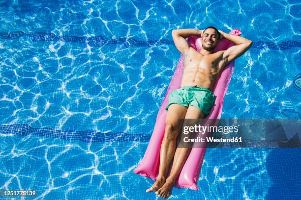 shirtless handsome young man with hands behind head relaxing on airbed in swimming pool - luftmatratze von oben stock-fotos und bilder