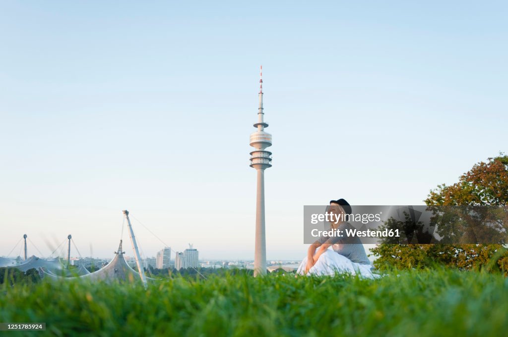 Thoughtful young woman sitting on grassy land with Olympic Tower in background at Munich, Germany