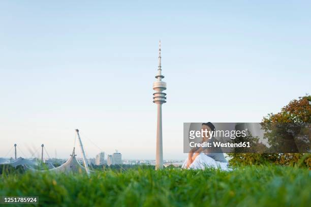 thoughtful young woman sitting on grassy land with olympic tower in background at munich, germany - olympiapark stock pictures, royalty-free photos & images