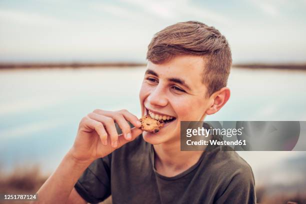 close-up of cheerful teenage boy eating cookie with lake and sky in background - eating cookies stock-fotos und bilder
